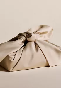 Nahaufnahme der Hand einer Person, die einen Knoten an einem beigen Furoshiki-Tuch bindet, vor einem beigen Hintergrund